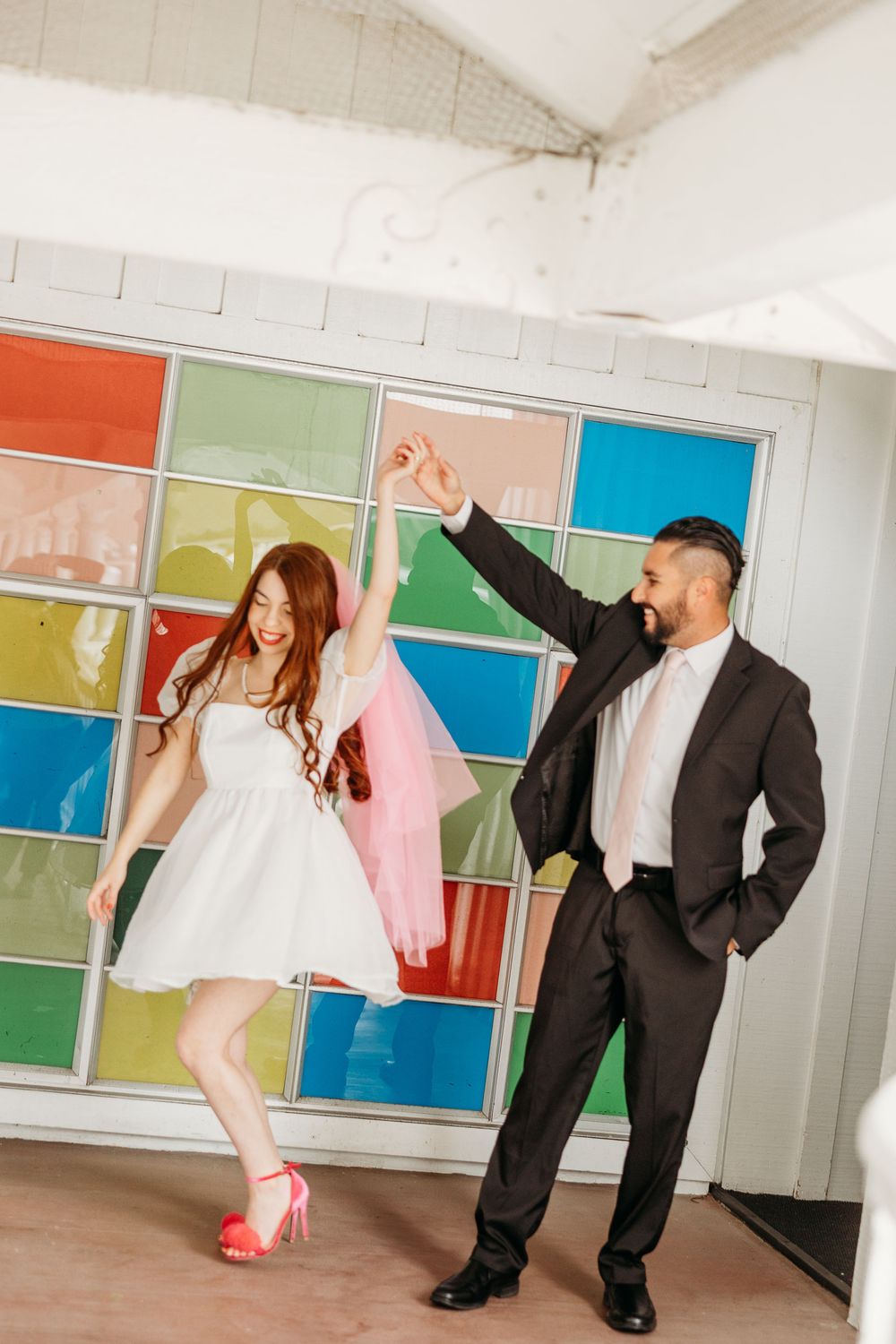 Two people dance joyfully in front of a colorful wall with geometric patterns at their wedding celebration.