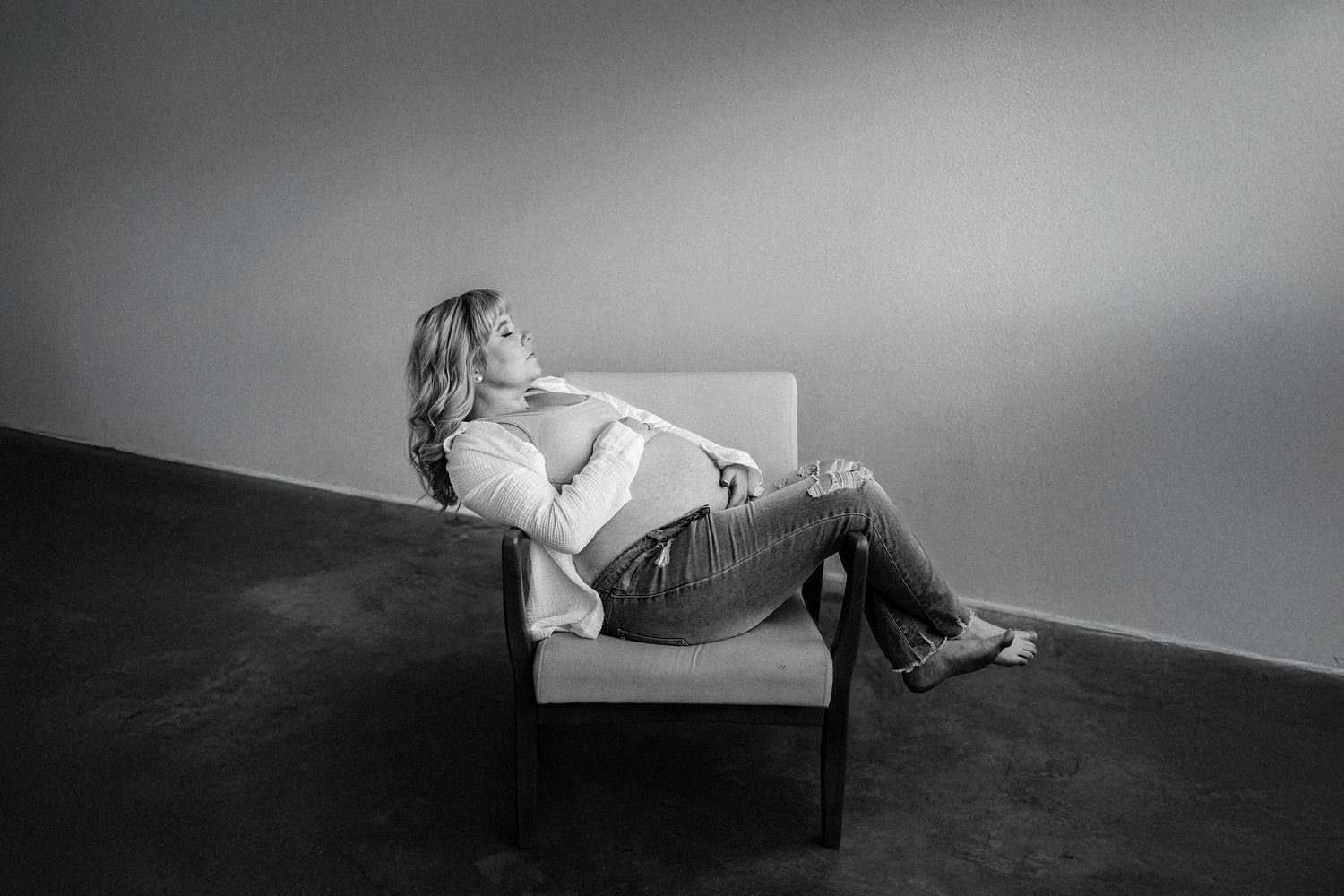 Pregnant woman in jeans and white shirt, relaxes on white chair against wall in dramatic black and white portrait.