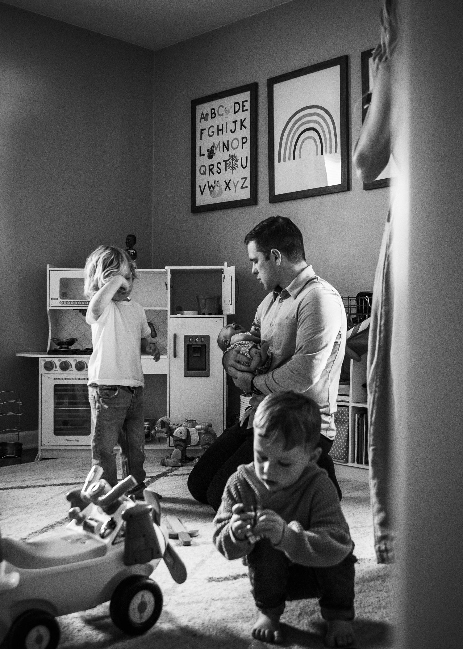 Black and white photo of a playroom with a toy kitchen set, framed wall art, and people playing with toys on the floor.