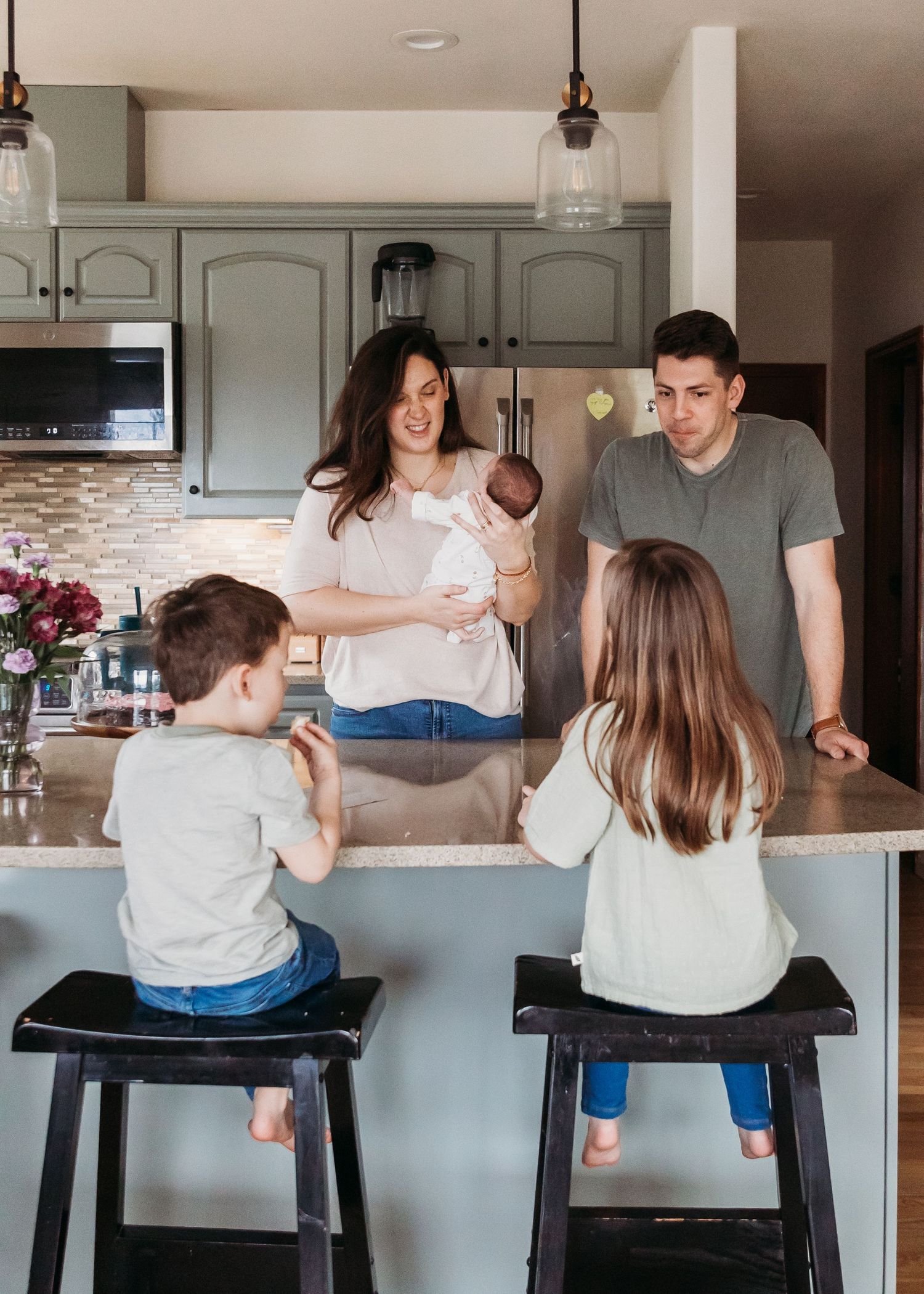 Family gathers around kitchen island counter with two children sitting on stools while parents stand and interact together.