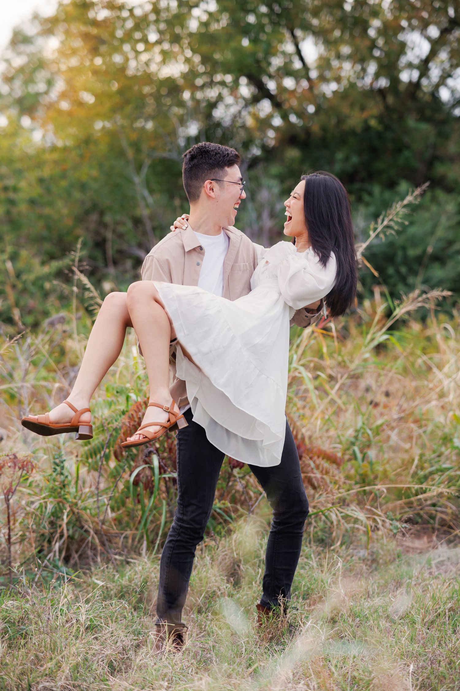 Couple in romantic outdoor engagement photoshoot with a man in beige sweater carrying a woman in white dress through grassy field.