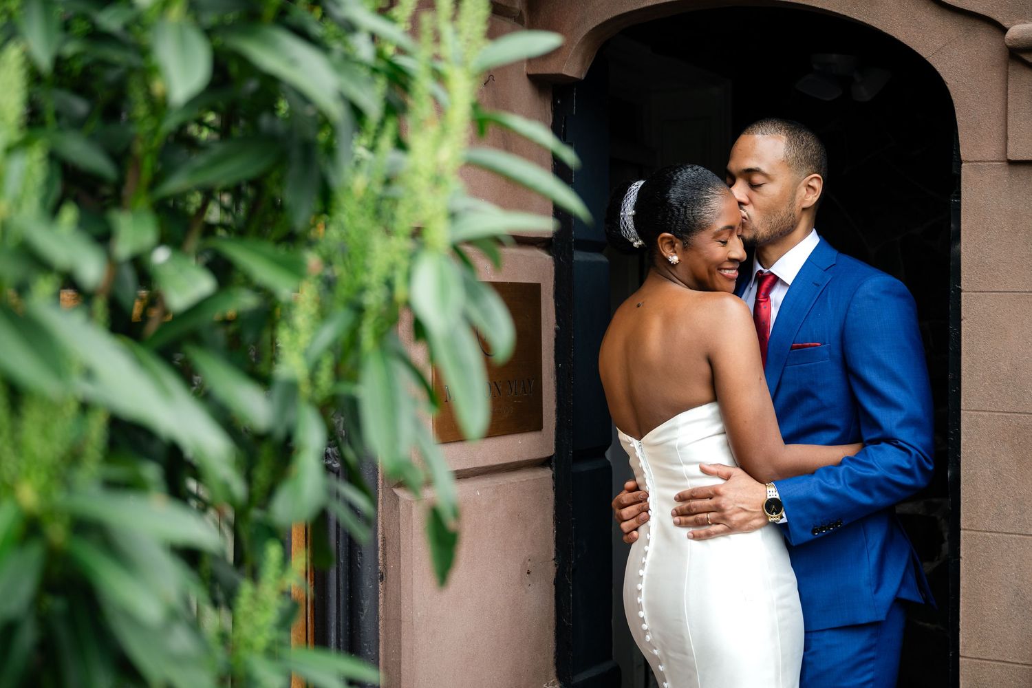 A wedding couple shares joyful moments on a brownstone staircase in an urban setting, wearing blue suit and white gown.