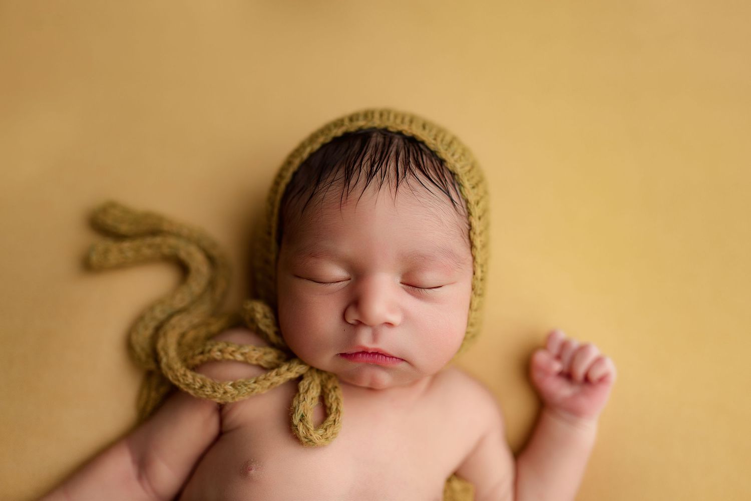 Sleeping newborn wearing a soft yellow knitted bonnet on neutral beige background.