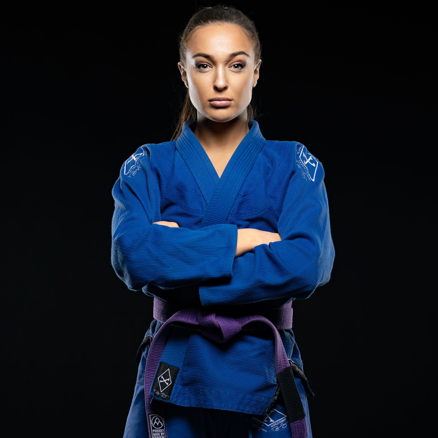 Kampsportutøver i blå judodrakt står selvsikkert med armene i kors mot en mørk bakgrunn.