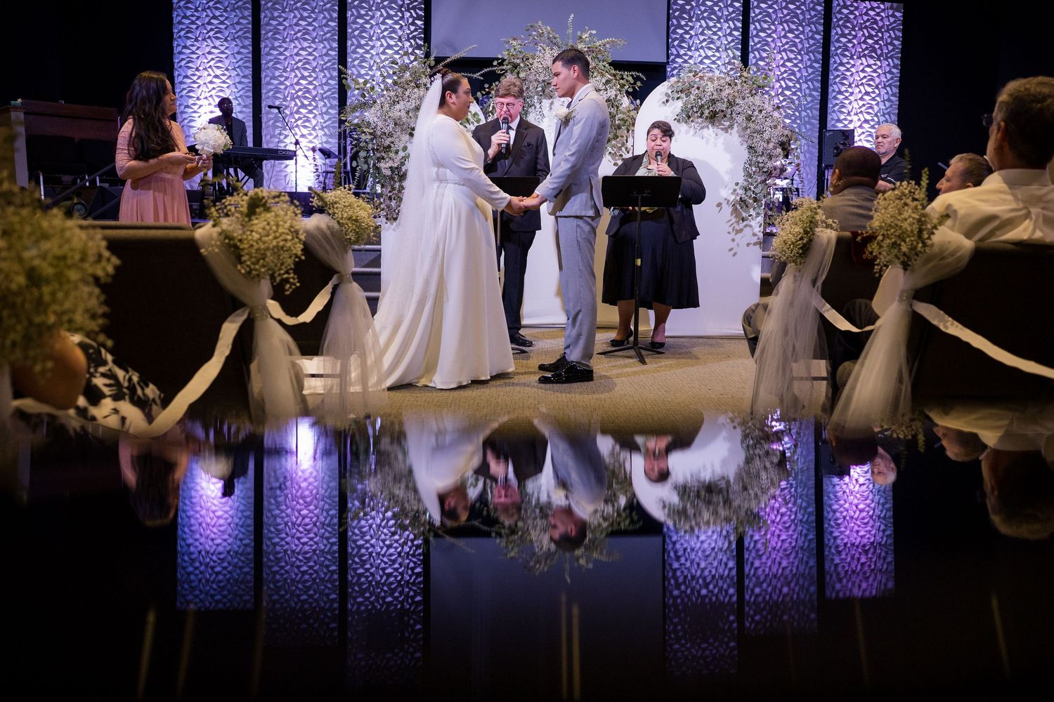 Wedding ceremony in progress on stage with purple uplighting and reflective floor creating mirror effect during evening service.