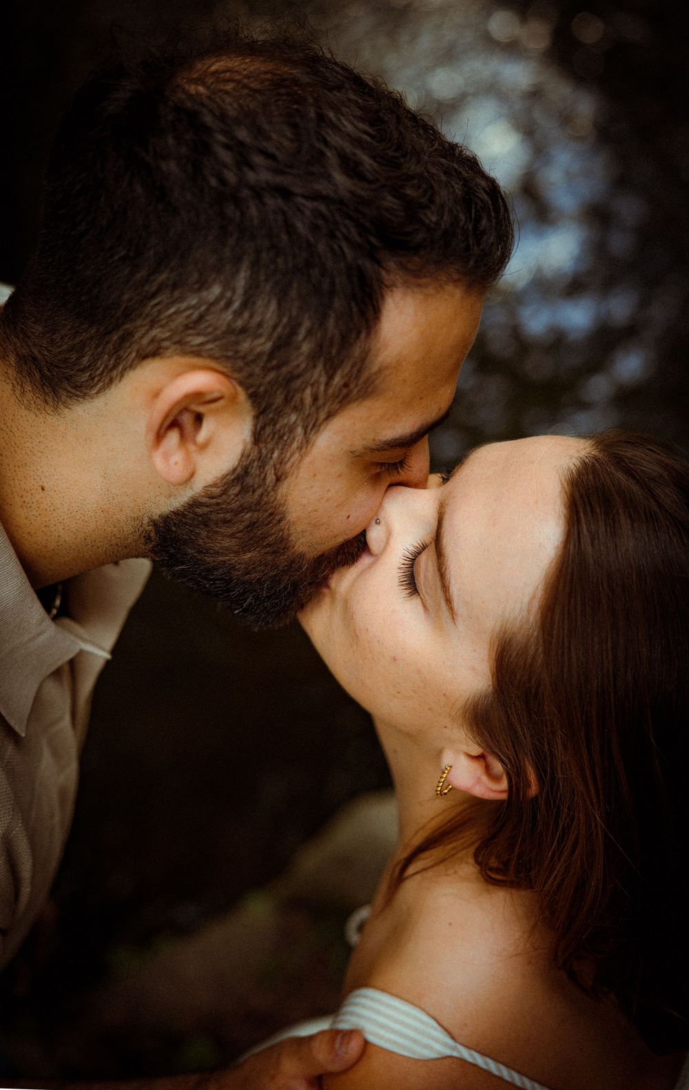 A romantic close-up photo of a couple sharing an intimate kiss outdoors with soft bokeh lighting in the background.