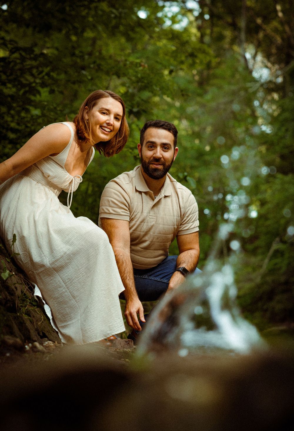 A couple poses together on a forest path with a soft blurred waterfall visible in the background.