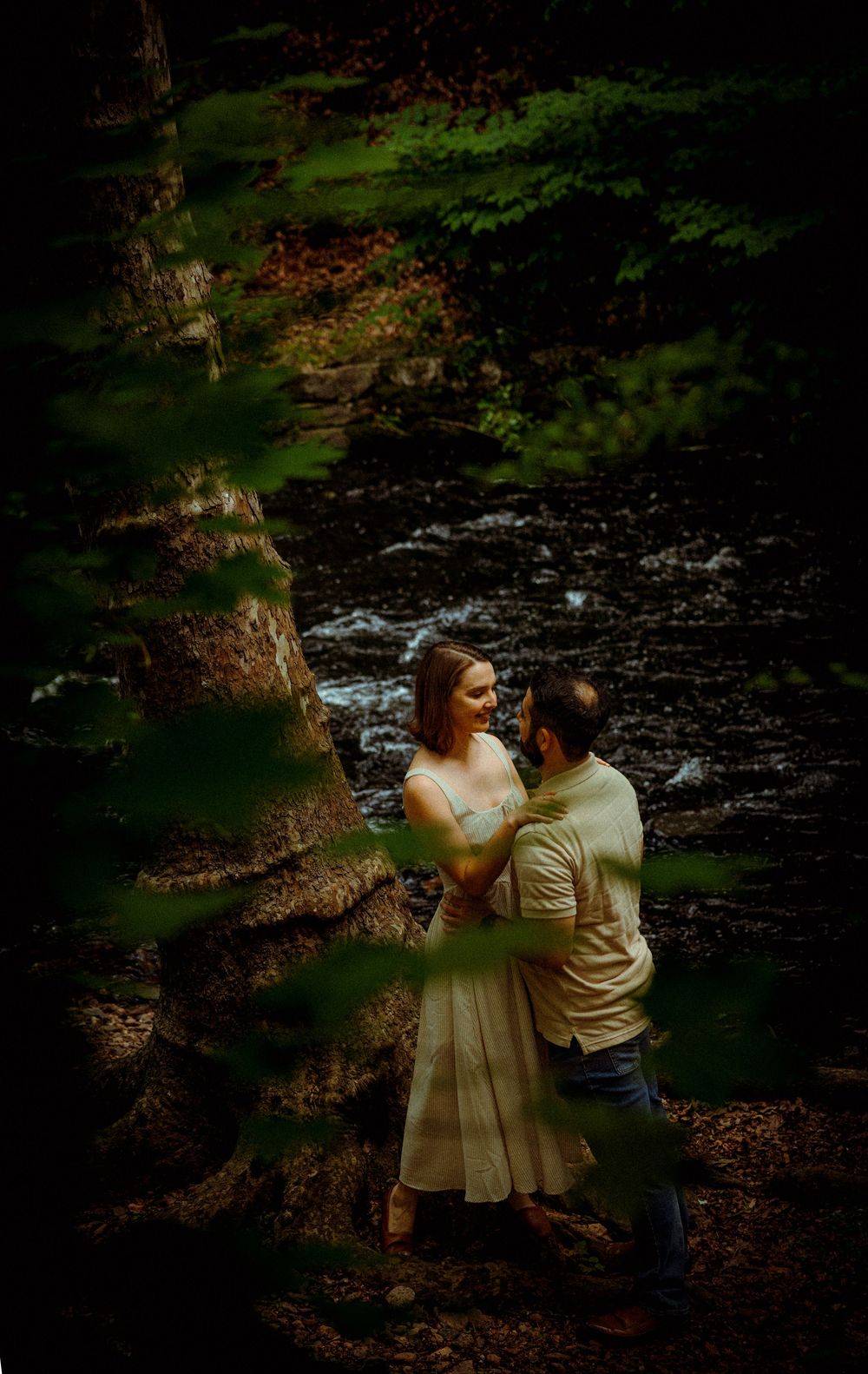 A romantic couple embraces beside a rushing stream in a lush forest setting with moody dark lighting.