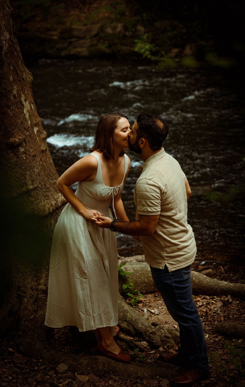A romantic moment captured by a rushing stream in a moody forest setting, with soft natural lighting and rustic scenery.
