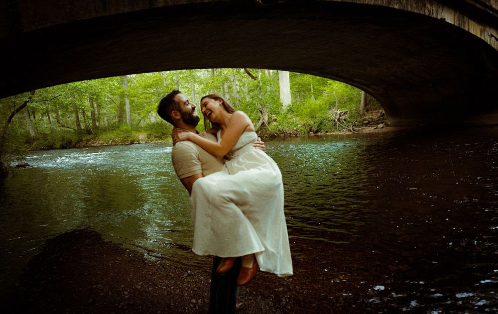 A romantic embrace under a stone bridge over a flowing stream in a lush green forest setting.