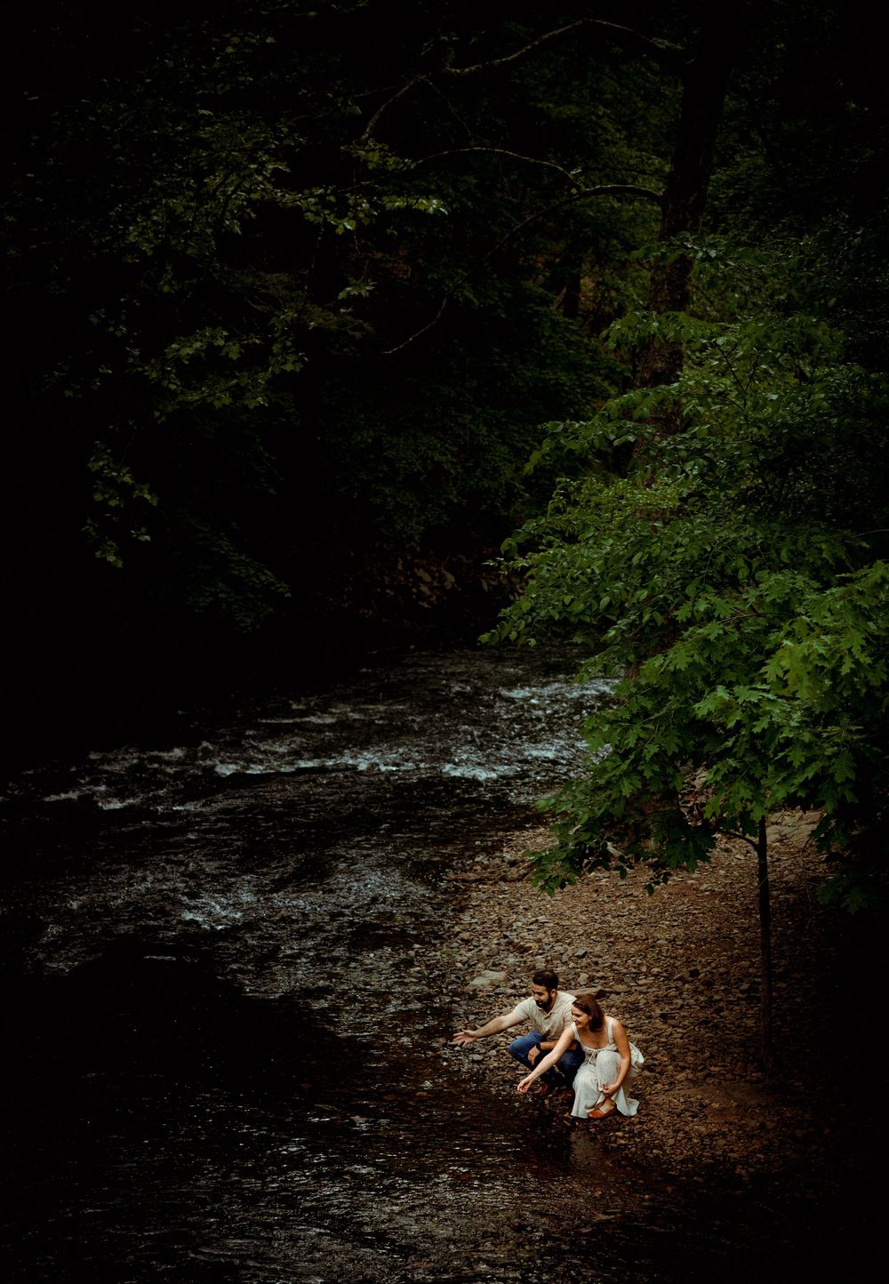 A couple shares a moment beside a flowing stream in a shadowy forest clearing.