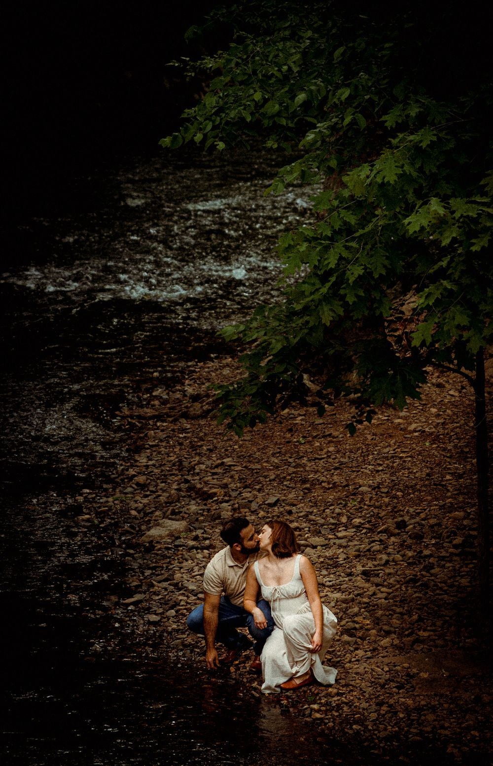 A romantic couple sits intimately by a stream in a lush forest setting with dramatic moody lighting.