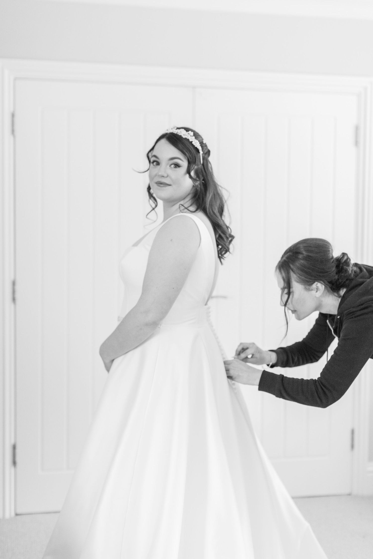 A bride in a white wedding dress has her gown fastened before the ceremony at a Cotswolds Village in Stow on the Wold - by Hayley Elizabeth