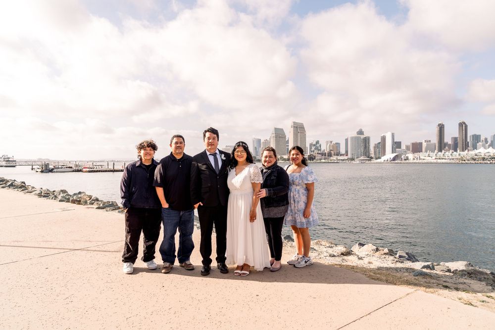 A group stands together on Coronado Island with the San Diego skyline and bay visible across the water on a cloudy day.