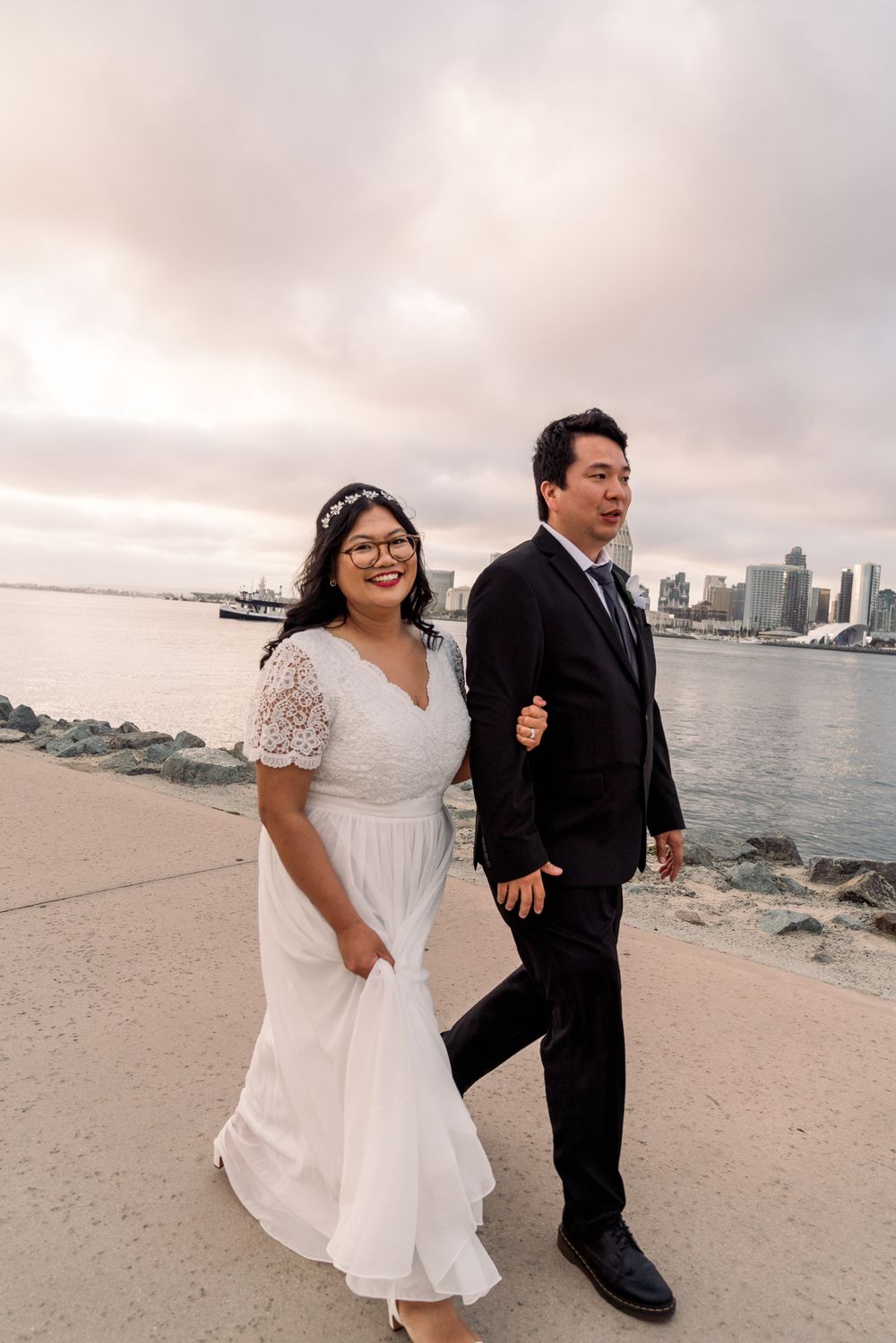A happy couple walks along the waterfront with a city skyline visible in the background on their wedding day.