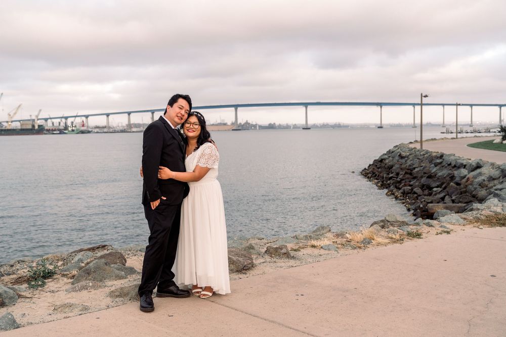 A couple embraces on a rocky shoreline with the Coronado Bridge spanning across San Diego Bay in the background.