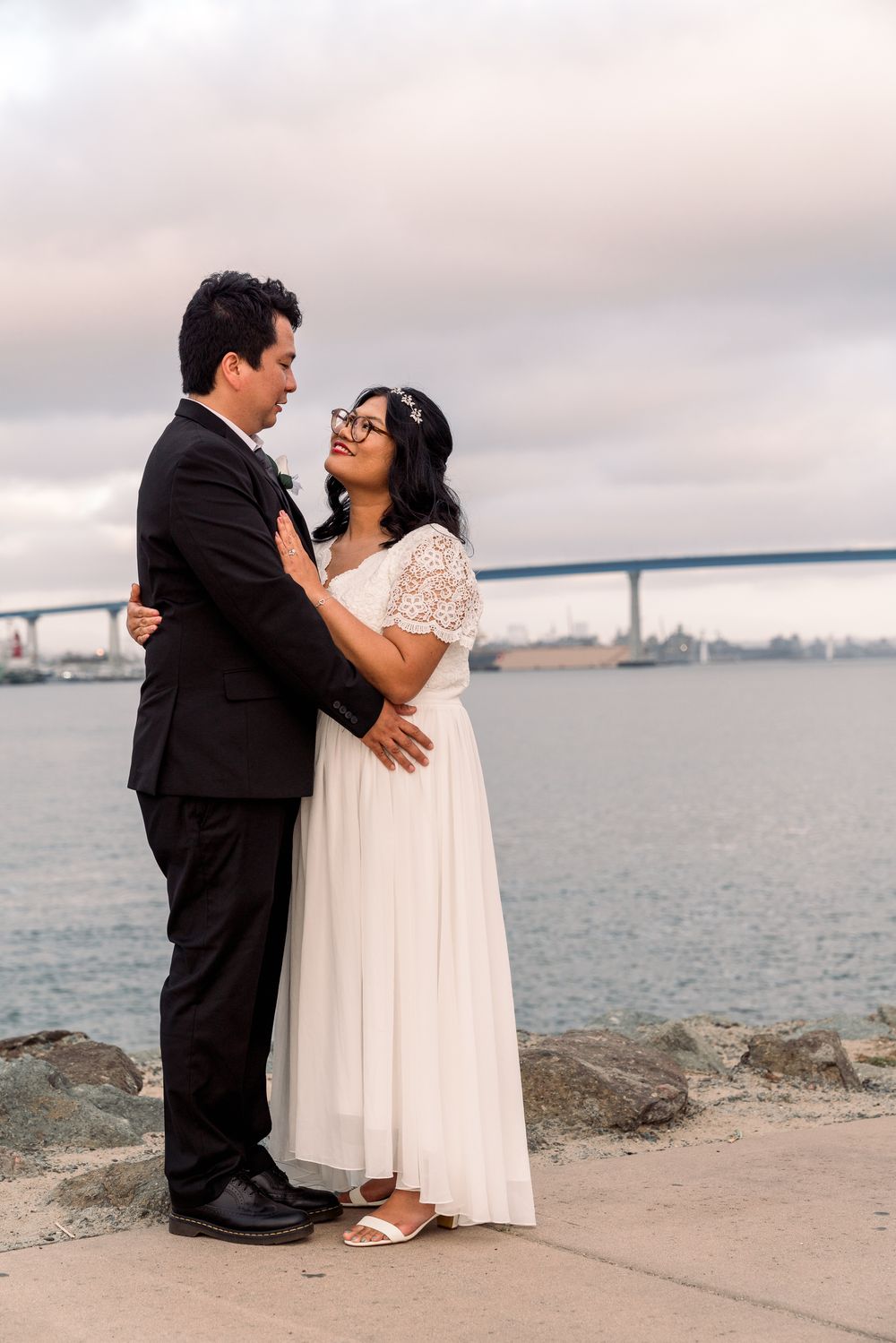 A couple in wedding attire stands on a rocky beach shore with the San Francisco Bay and bridge in the distance.