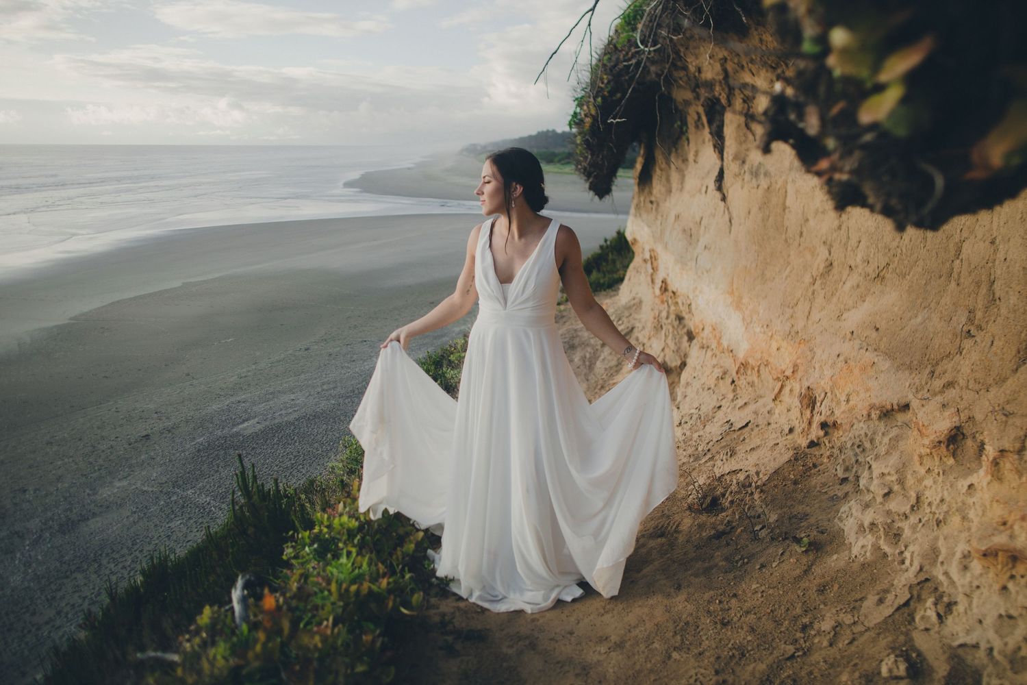 A bride in a flowing white dress stands elegantly on a coastal cliff overlooking a misty ocean view.