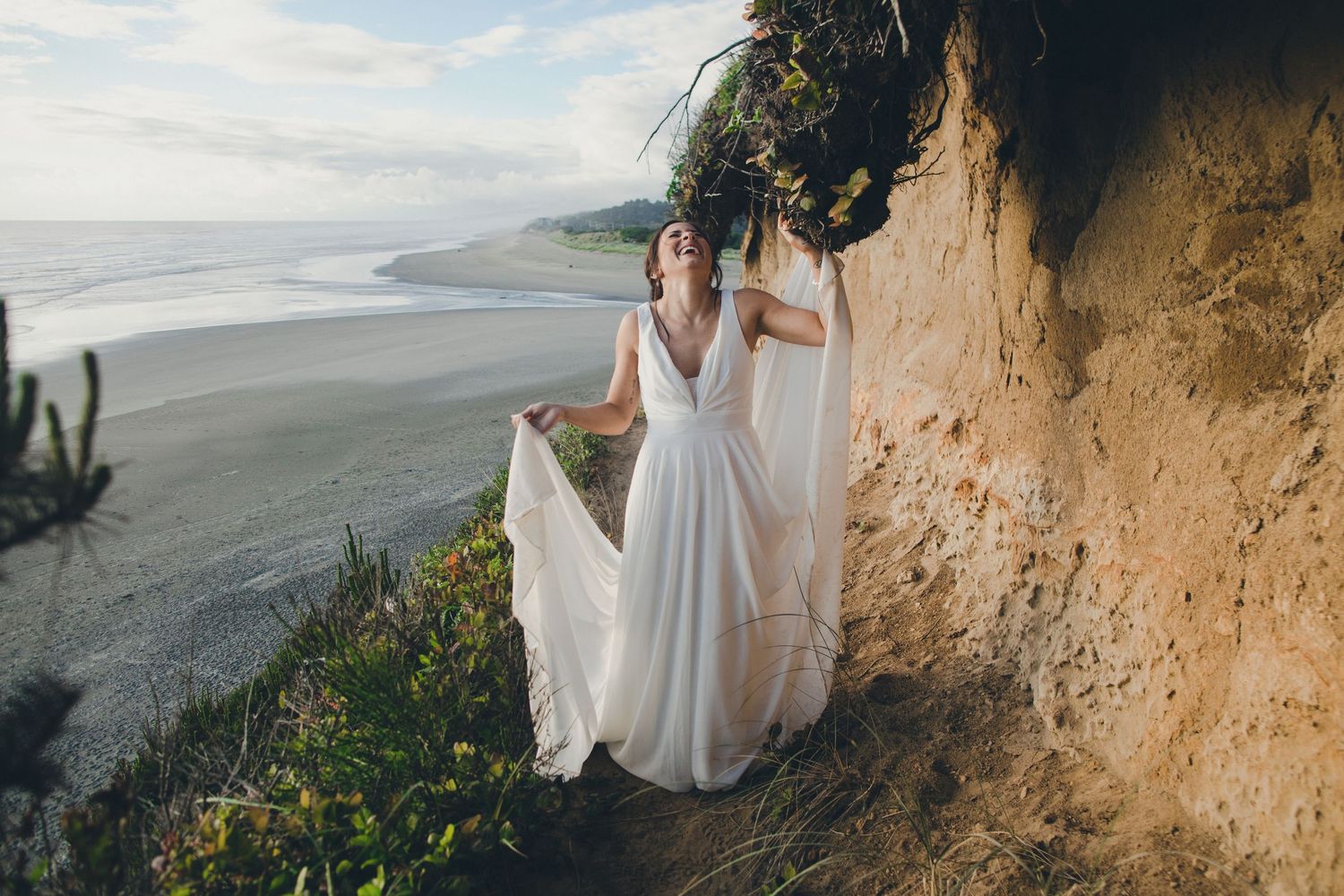 Two people in flowing white dresses stand on a dramatic coastal cliff overlooking a misty beach landscape.