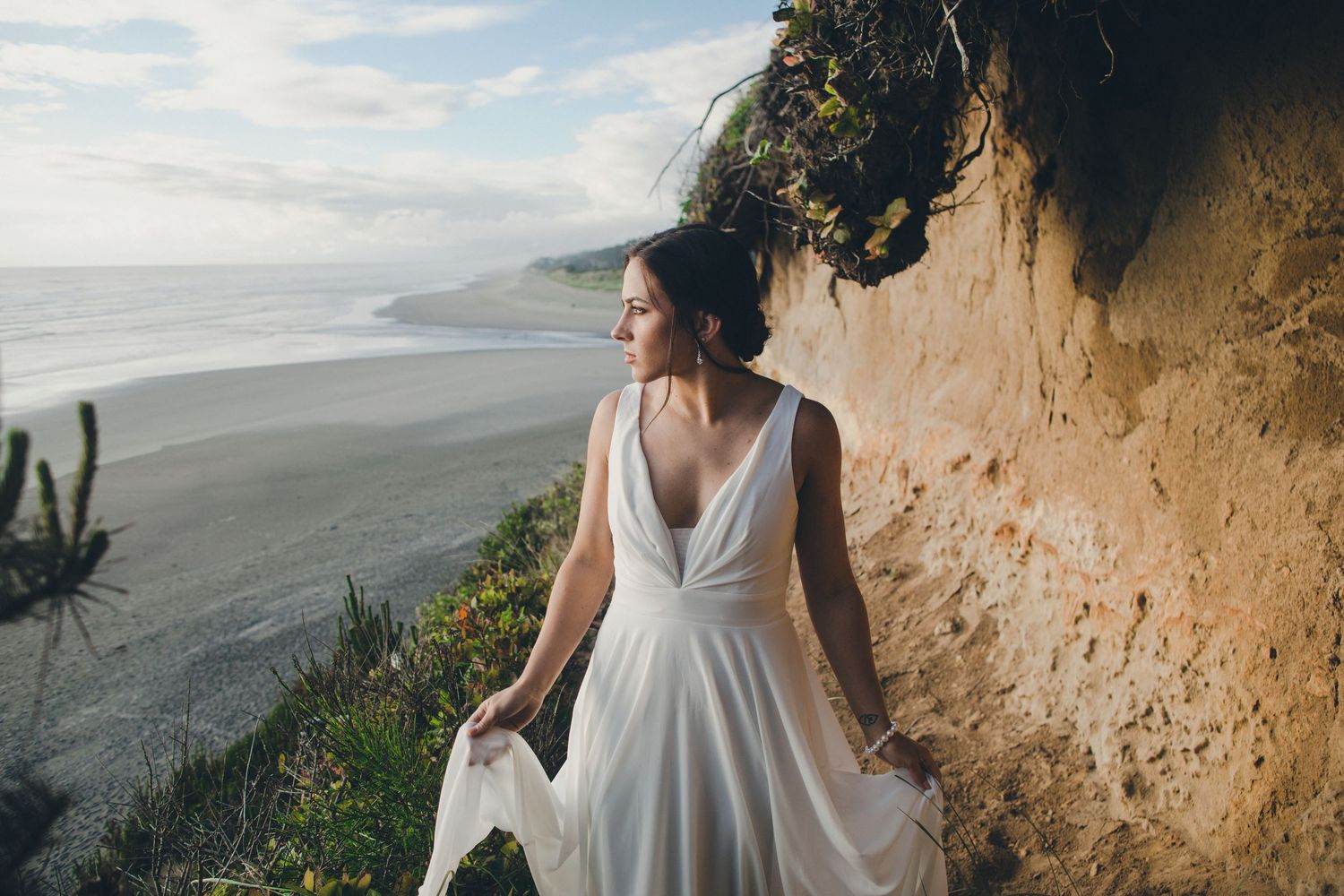 A bride in a white flowing dress stands against a coastal cliff wall overlooking a misty beach landscape.