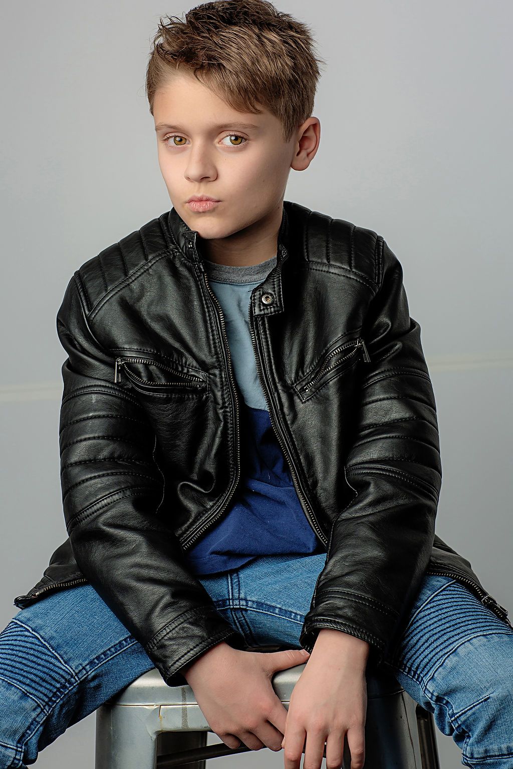 Young person wearing a black leather jacket and blue jeans sitting on a stool against a light gray background.