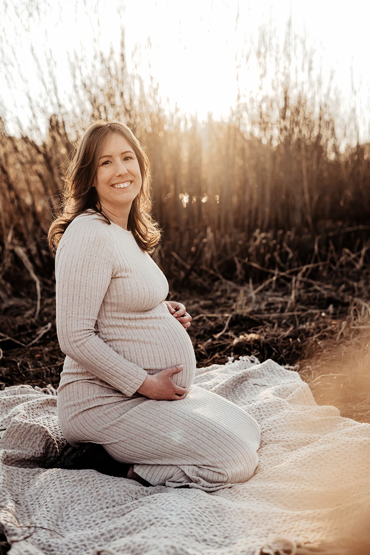 Gravidfotografering, en gravid kvinna sitter på en filt i varmt kvällsljus som silar genom vassen vid strandkanten, hon ler in i kameran.