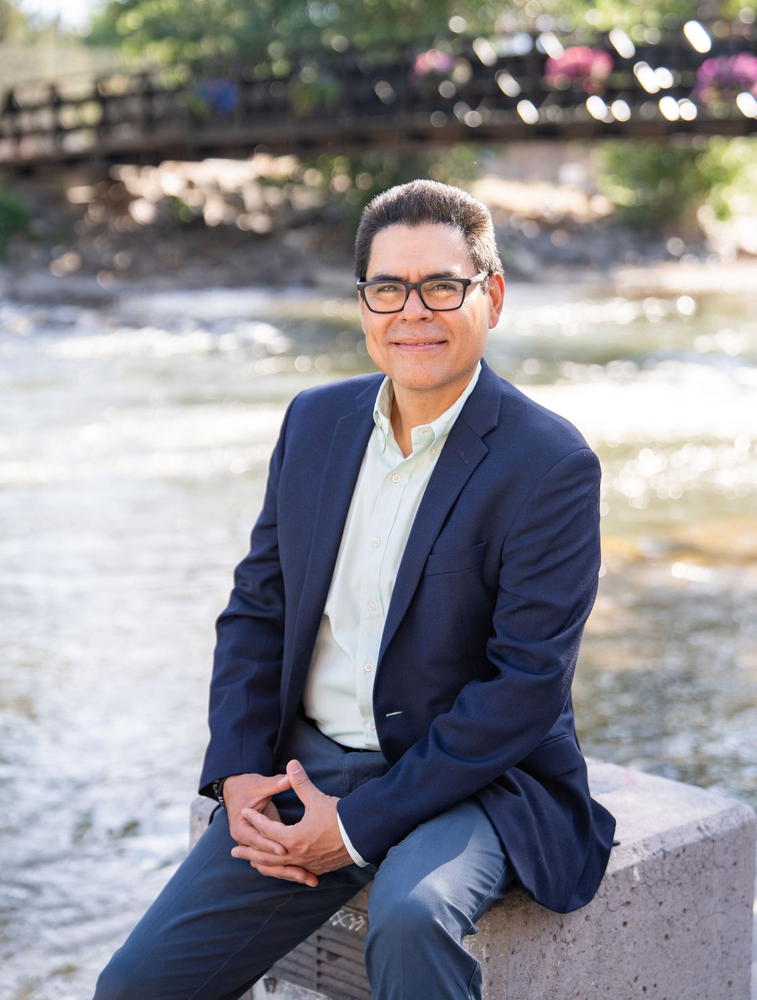 Professional wearing a navy blazer and white button-down shirt poses outdoors against a lush green background.
