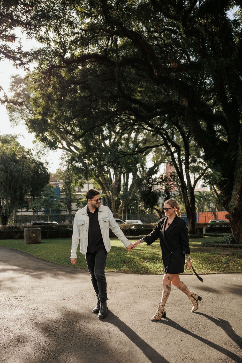 A couple holds hands while walking along a tree-lined path in a park during golden hour.
