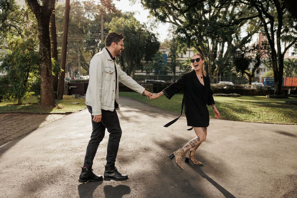 Two people playfully hold hands while balancing on a concrete path in a sunny park setting.