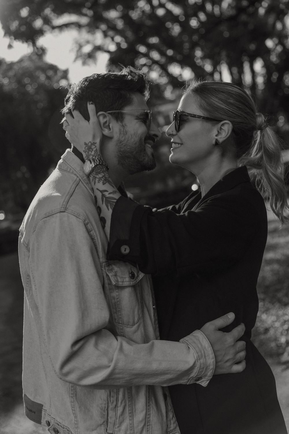 A romantic black and white portrait of a couple in sunglasses sharing an intimate moment outdoors at sunset.