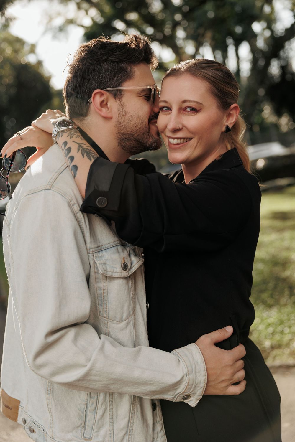 A loving couple embraces outdoors on a sunny day, with one wearing a light denim jacket and the other in black.