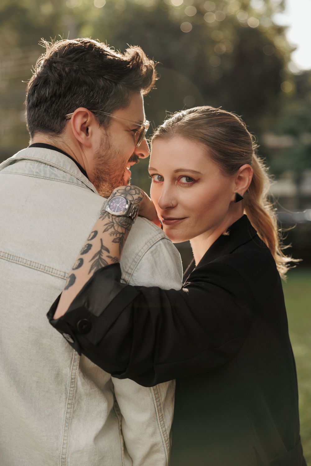 Romantic outdoor portrait of a couple embracing, with person in black top smiling at camera and partner wearing denim jacket.