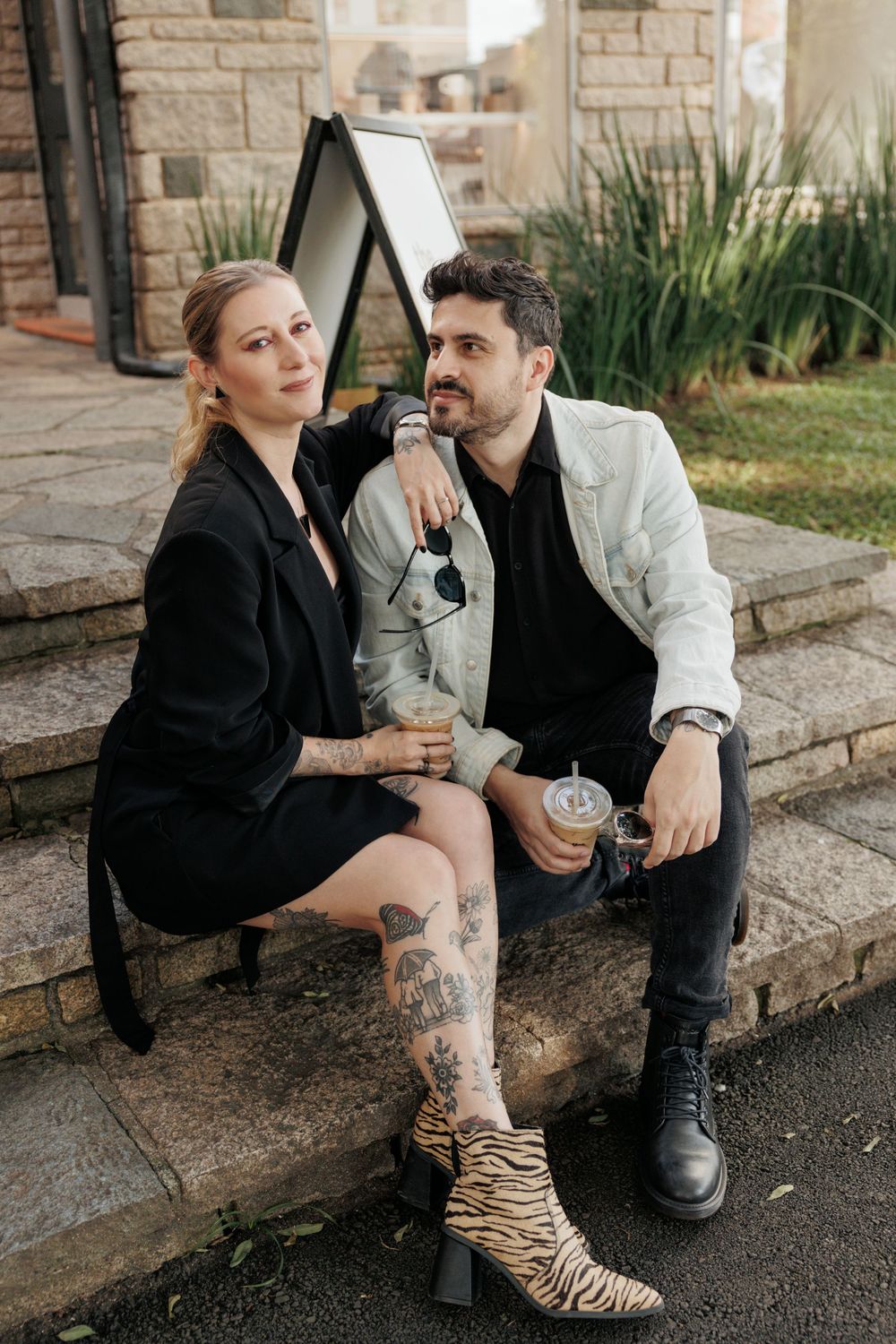 A stylish couple sits on stone steps, one in a black dress and tiger boots, the other in a white blazer holding drinks.