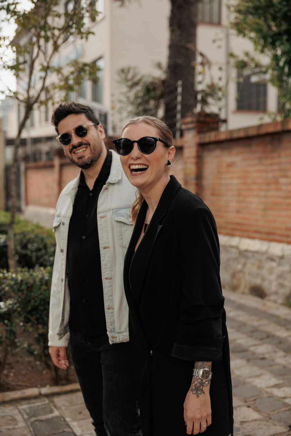 Two friends in sunglasses and dark jackets share a laugh together on a street with brick walls and trees in the background.