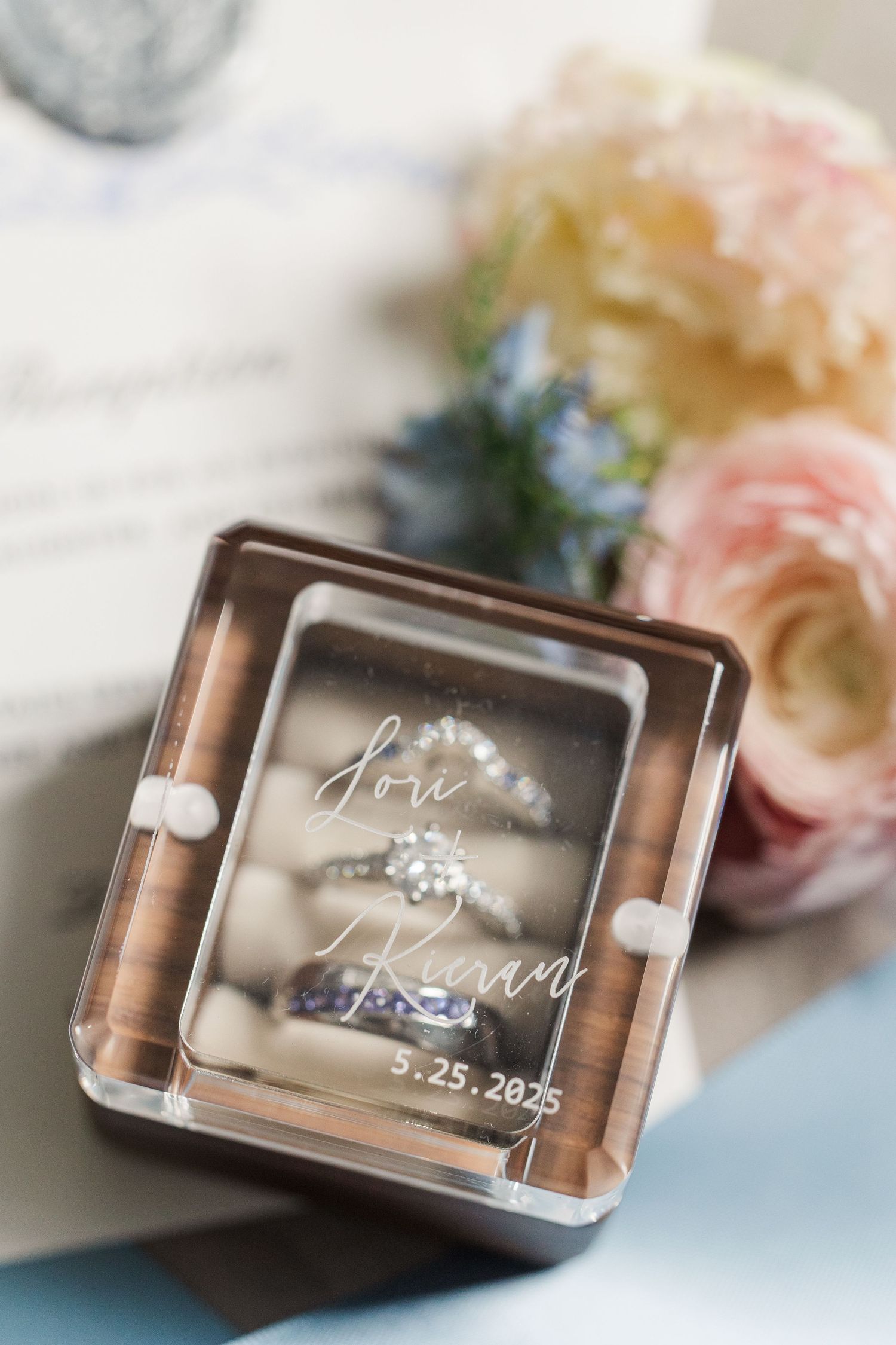 Engagement ring displayed in clear acrylic box with wedding date 2.25.2025 and pink roses in soft focus background.