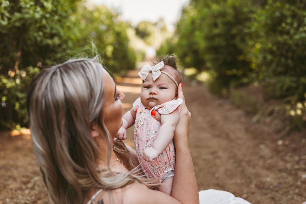 Parent holds and gazes lovingly at baby wearing floral headband in sunlit orchard path.