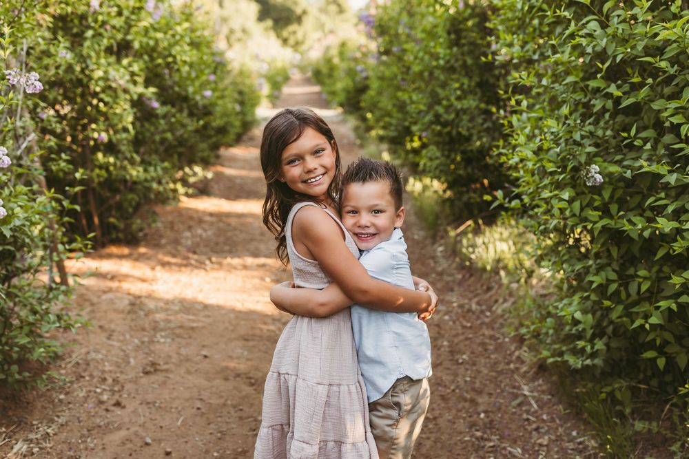 Two children embrace and smile while standing on a dirt path surrounded by green bushes in warm sunlight.