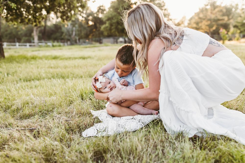 Someone wearing a white dress leans over tenderly while sitting on a blanket with a baby in a grassy field.