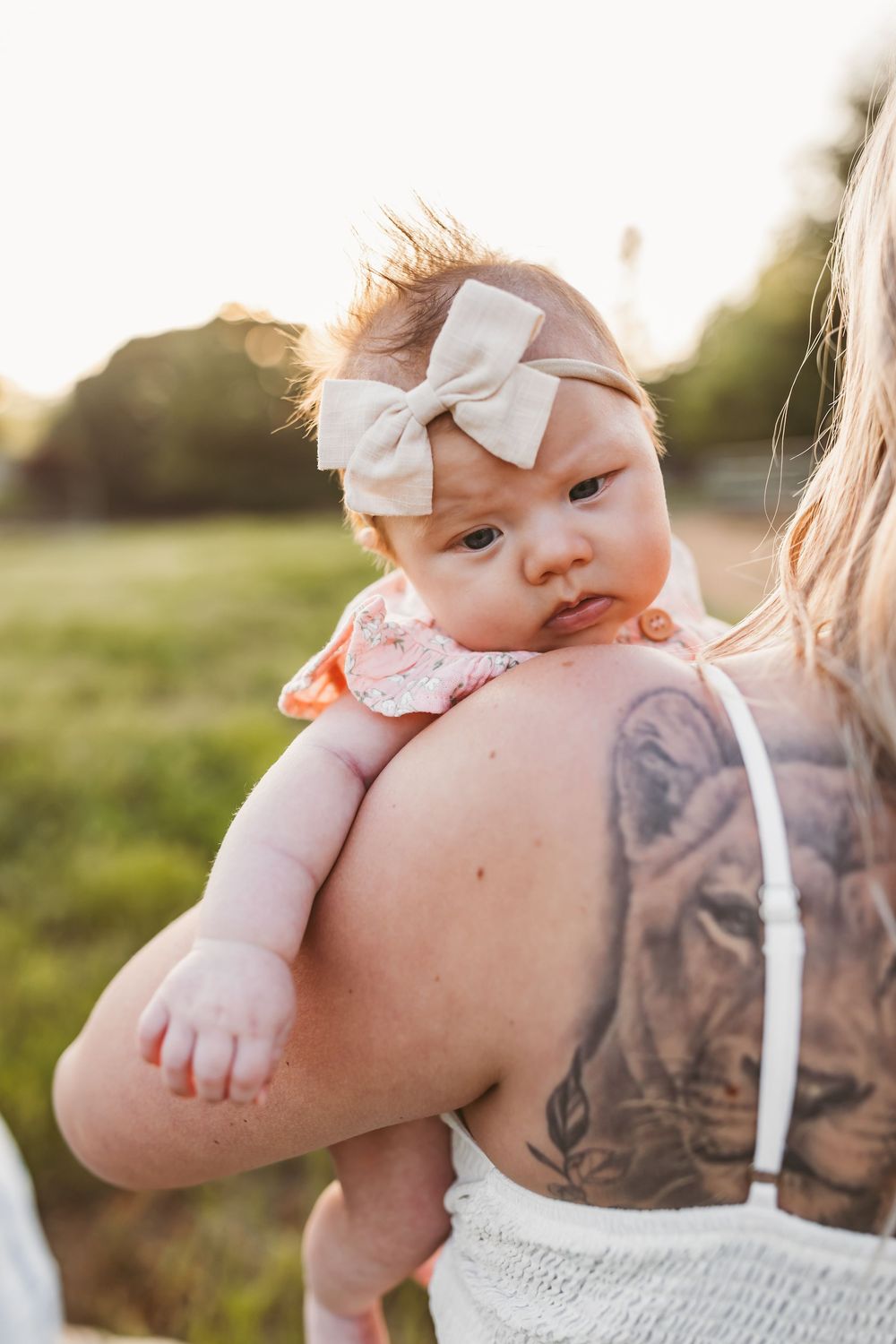 A small baby wearing a white bow headband is held against someone with a visible floral tattoo at sunset in a field.