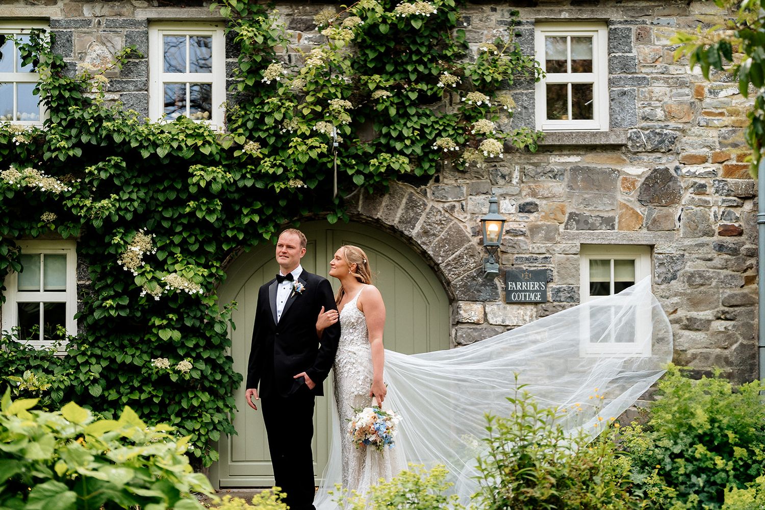 Elegant couple poses in front of a stone house with ivy-covered walls and a beautiful flowing veil catching the breeze.