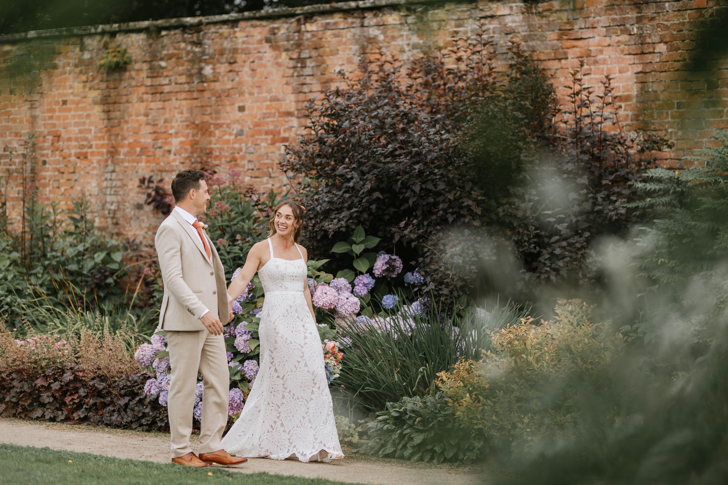 Two people in wedding attire stand together in a garden with an old brick wall and purple flowers in the background.