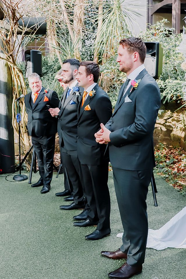 Groomsmen in matching dark suits stand in line on green carpet during an outdoor garden wedding ceremony.