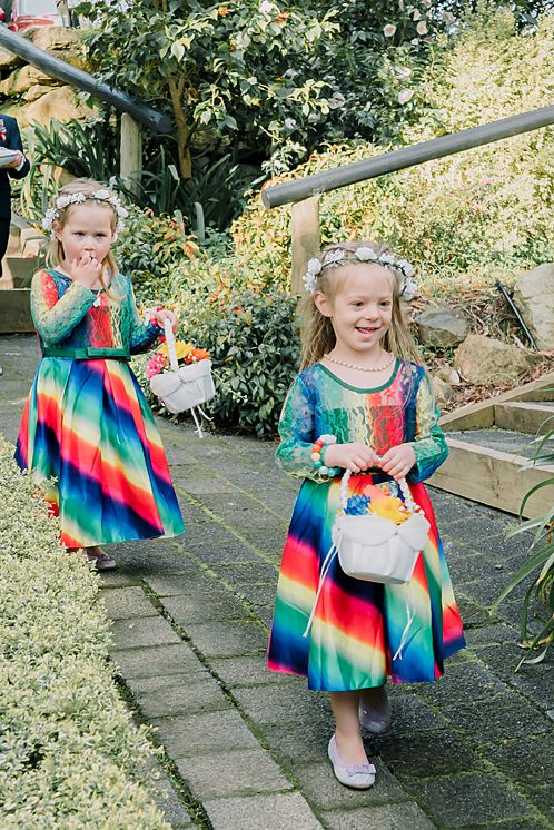 Two children in vibrant rainbow-striped dresses carry white baskets while walking along a garden path.