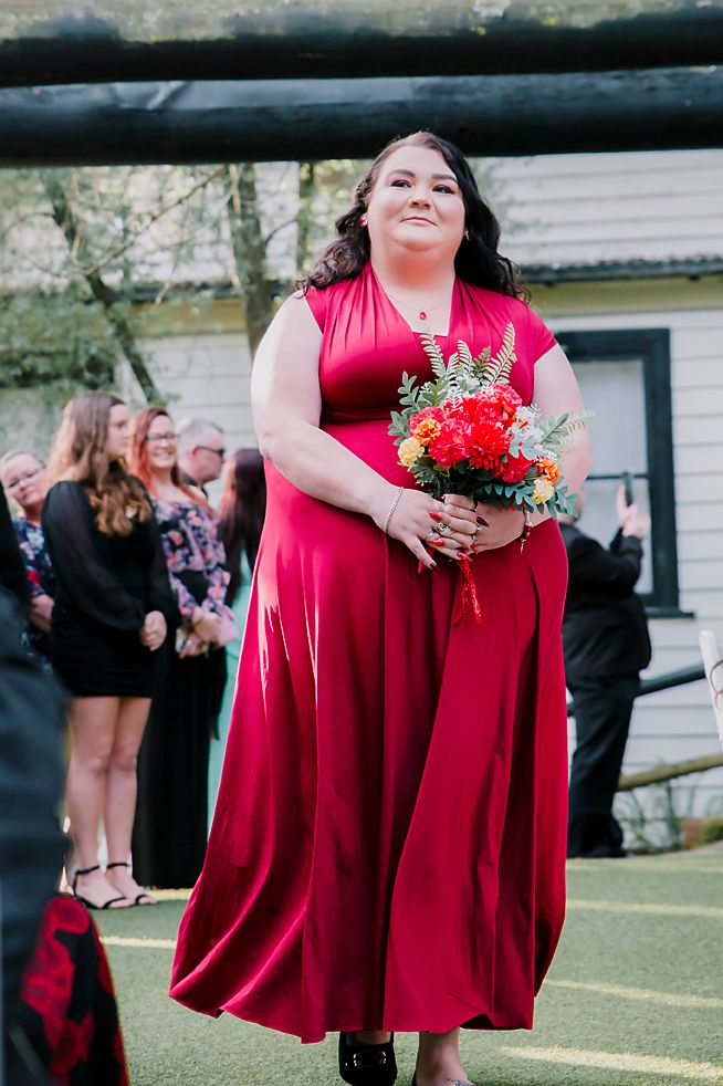 Wedding party member in flowing red dress holds vibrant floral bouquet during outdoor celebration.