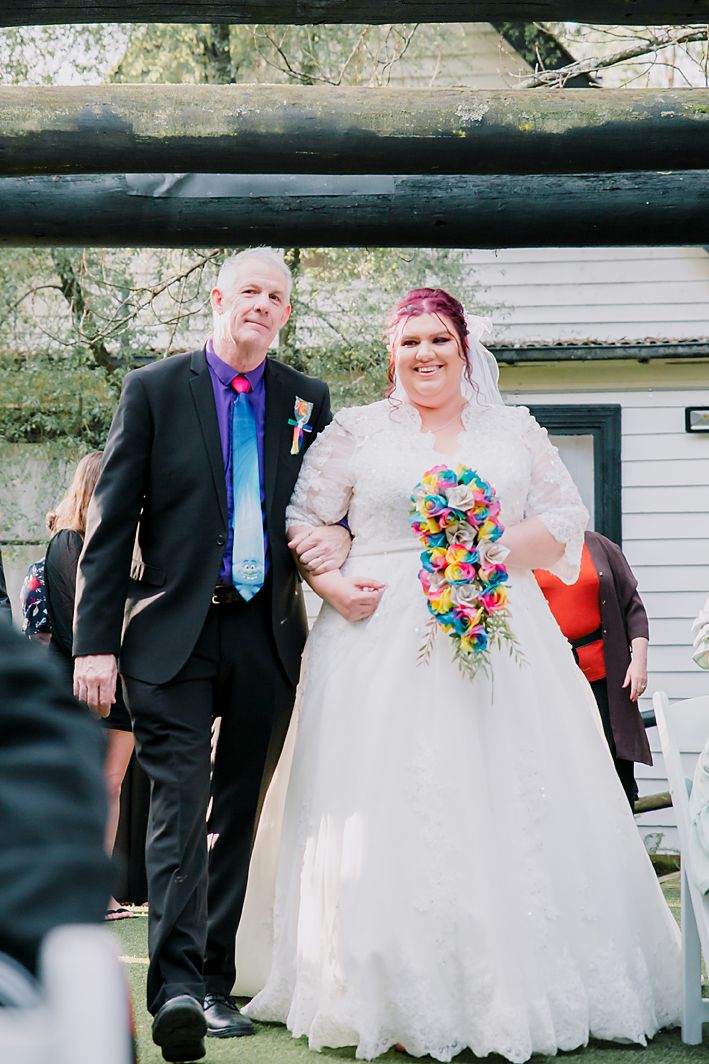 Wedding entrance with colorful cascading tropical bouquet and formal attire against a white building backdrop.