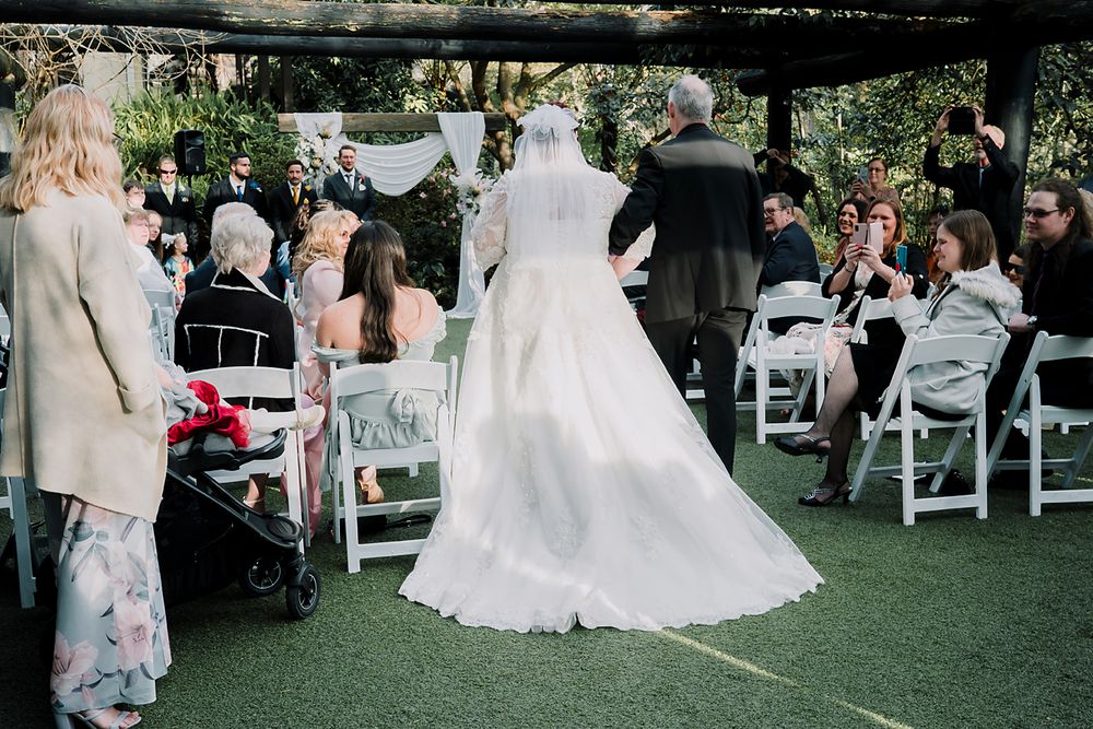 Wedding ceremony taking place outdoors on green turf with white chairs and flowy white wedding dress in view.