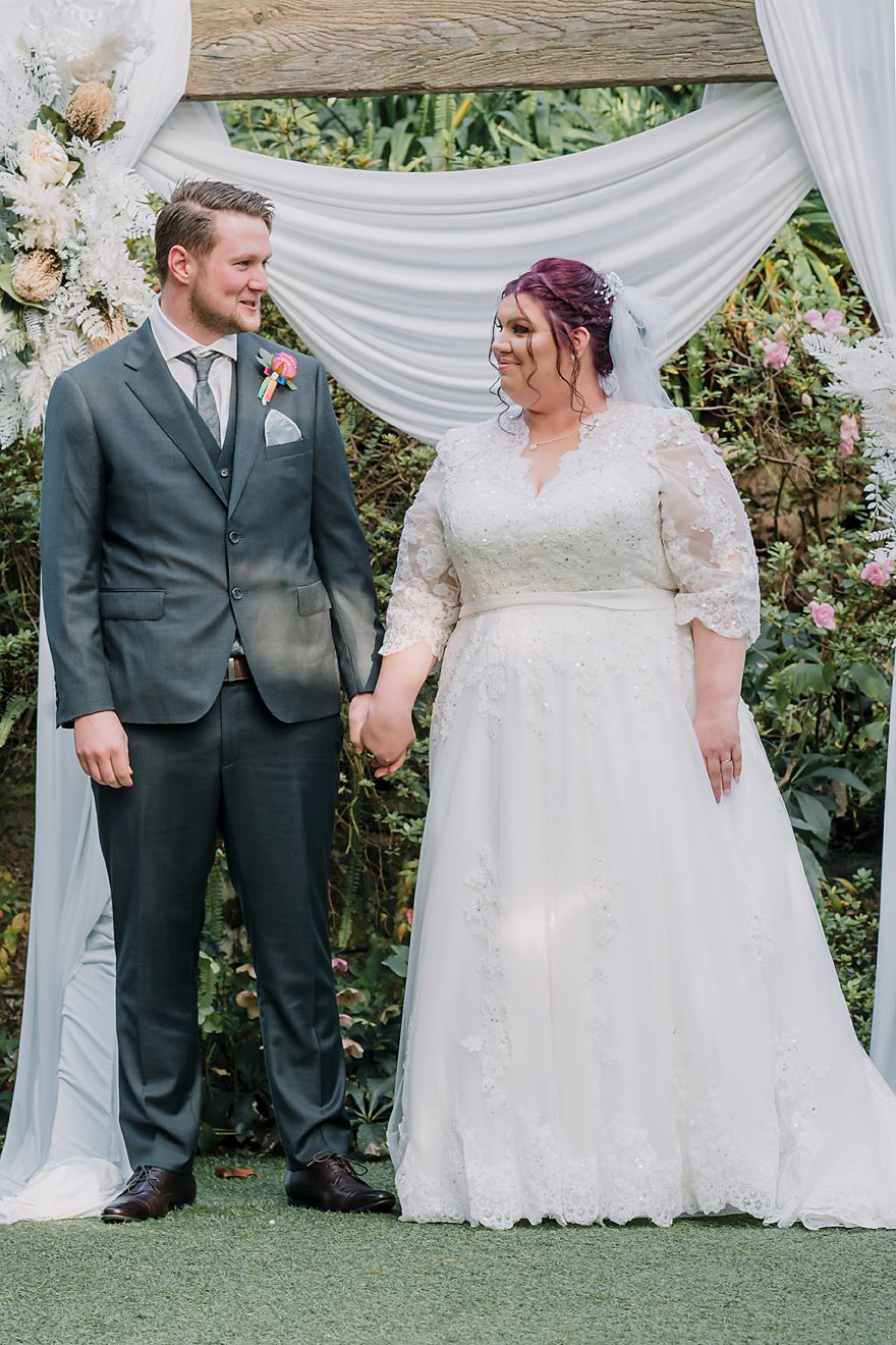 Couple wearing a white wedding gown and grey suit pose together against white draping and greenery backdrop.