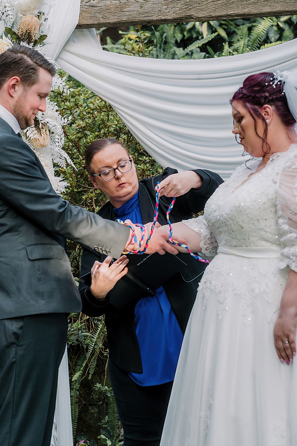 Wedding couple rosary/handfasting ceremony with officiant in royal blue during outdoor garden ceremony.