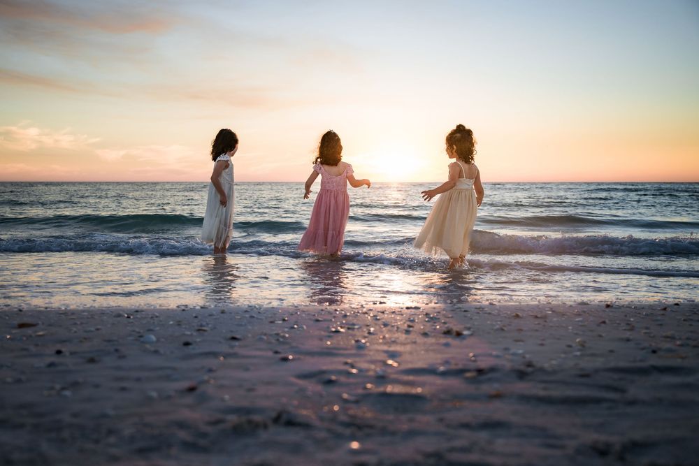 Three silhouettes of people in flowing dresses walk along a sunset beach with waves crashing on the shore.