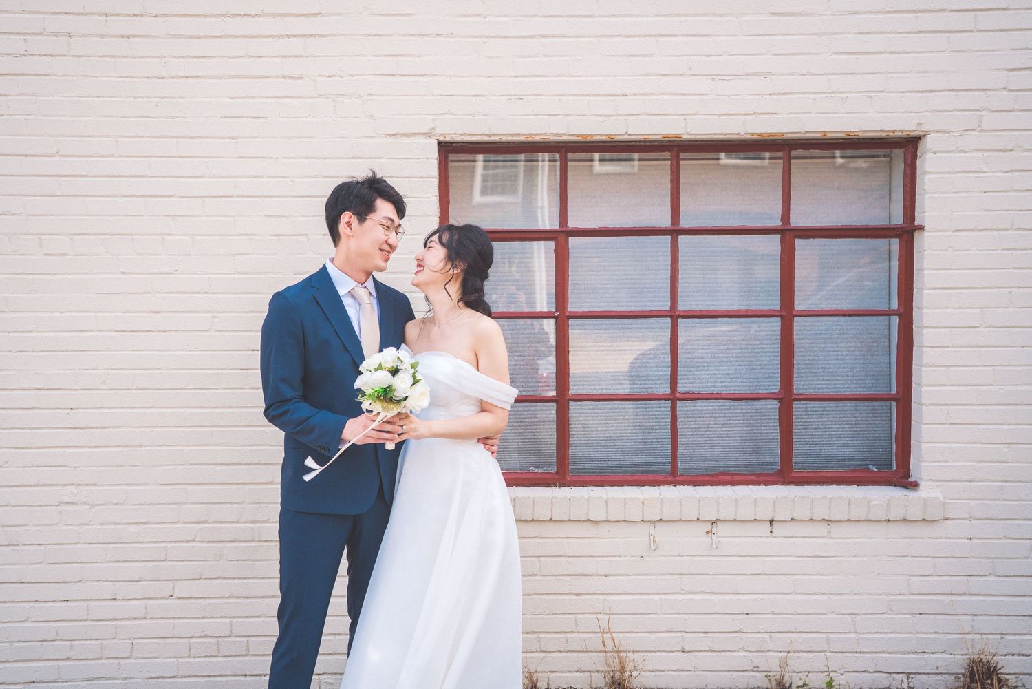 A couple in wedding attire smiles at each other while holding a flower bouquet against a beige brick wall with a window.